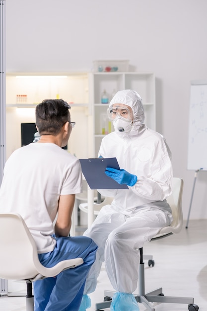 Química ou cientista feminina contemporânea em roupas de trabalho de proteção, consultando um homem chinês em um hospital ou laboratório