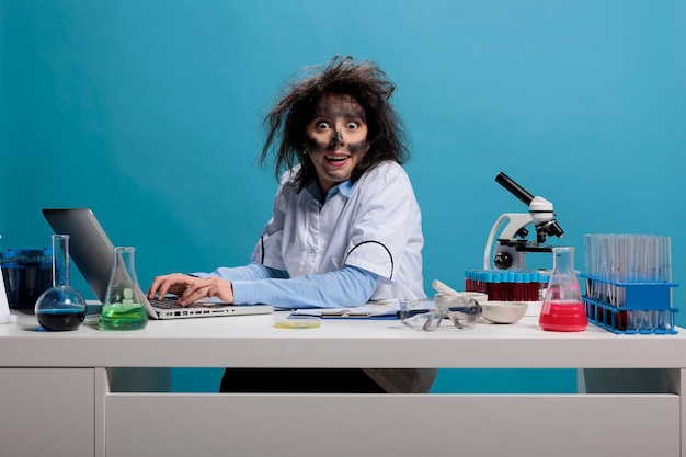 Químico bobo louco com olhar selvagem sentado na mesa trabalhando no laptop enquanto sorrindo assustador para a câmera no fundo azul. Trabalhador de laboratório insano com aparência esfumaçada após explosão química tóxica