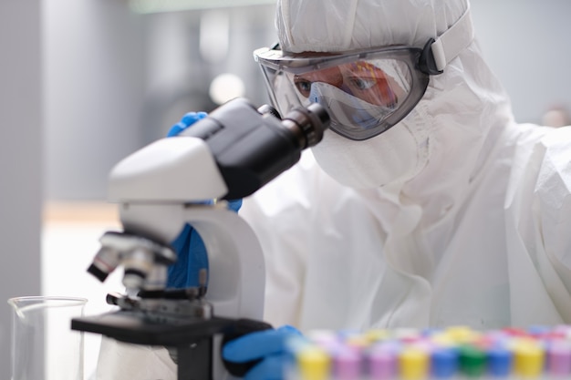 Químico científico en traje de protección antiplaga mirando a través del microscopio en el laboratorio