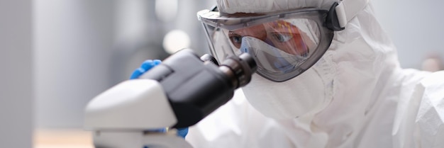 Químico científico en traje protector antiplaga mirando a través del microscopio en el laboratorio