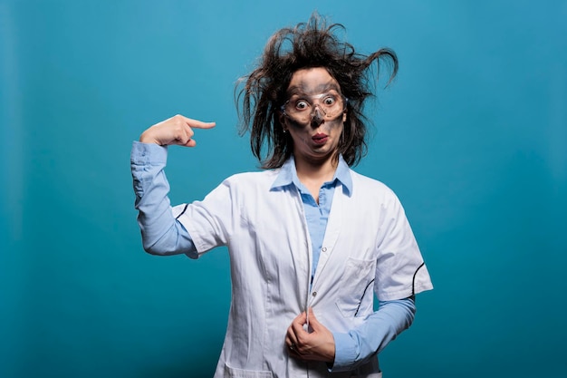 Químico loco y de aspecto divertido que gesticula locura torciendo el dedo en la sien mientras tiene la cara sucia y el cabello desordenado. Científico divertido con apariencia extravagante mirando a la cámara sobre fondo azul.