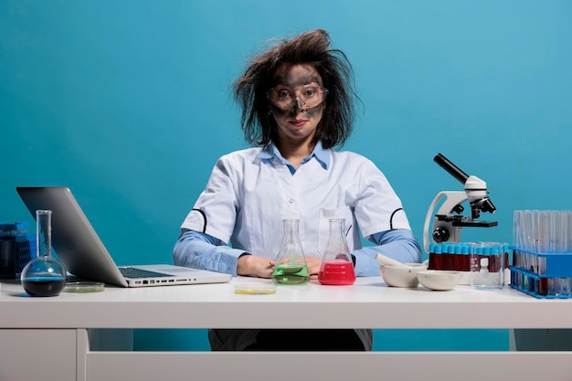 Químico louco de aparência boba com cabelo bagunçado e rosto sujo sentado na mesa após a explosão do experimento de laboratório fracassado. Trabalhador de laboratório tolo com aparência selvagem sentado sobre fundo azul. Foto de estúdio