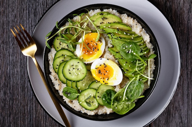 Quinoa-Schale mit Eierspitze mit Sonnenblumen-Mikrogrün, Gurke, Avocado, Babyspinat in einer schwarzen Schüssel auf einem dunklen Holztisch, flache Lage, Nahaufnahme, türkische Küche