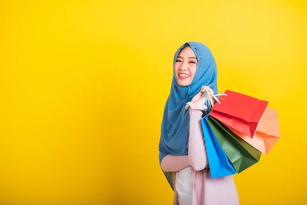 Árabe muçulmano asiático, retrato de uma jovem bonita e feliz Islã desgaste religioso véu hijab sorriso engraçado ela segurando sacolas de compras coloridas, tiro de estúdio isolado em fundo amarelo