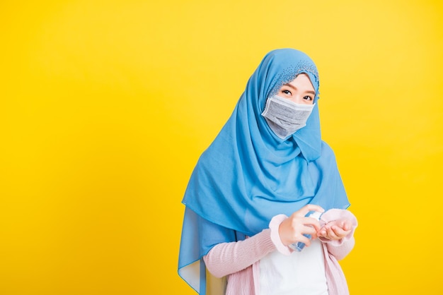 Árabe muçulmano asiático, retrato de uma jovem bonita e feliz, o uso religioso do Islã, o véu hijab e a máscara facial protegem a doença do coronavírus em quarentena, aplique spray de álcool nas mãos isoladas em amarelo