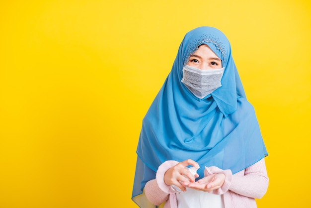Árabe muçulmano asiático, retrato de uma jovem bonita e feliz, o uso religioso do Islã, o véu hijab e a máscara facial protegem a doença do coronavírus em quarentena, aplique spray de álcool nas mãos isoladas em amarelo