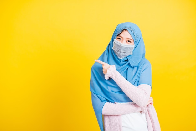 Árabe muçulmano asiático, retrato de uma jovem bonita e feliz religiosa usa véu hijab e máscara protetora para prevenir o coronavírus, ela aponta o dedo para o lado, isolado em fundo amarelo