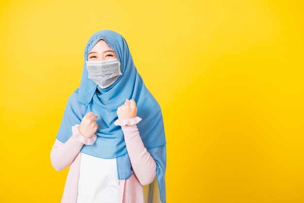 Árabe muçulmano, jovem bonita e feliz usa véu hijab usa máscara facial protetora quarentena coronavírus levanta as mãos feliz animado alegre depois de se recuperar de doença, fundo amarelo isolado