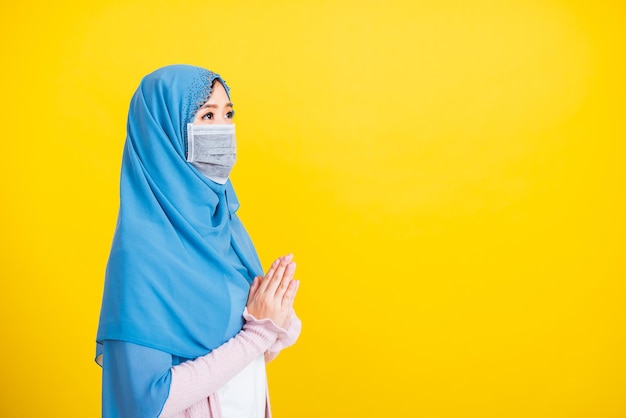 Árabe musulmán asiático, Retrato de una hermosa joven religiosa islámica usa velo hijab y máscara facial protege las cuarentenas enfermedad coronavirus levantando la mano rezando Eid al Fitr fondo amarillo aislado