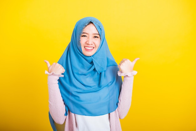 Árabe musulmán asiático, Retrato de una joven feliz y hermosa, religiosa islámica, con velo hijab, sonrisa graciosa que hizo con los dedos pulgares hacia arriba, señal de acuerdo para estar de acuerdo, toma de estudio aislada en fondo amarillo