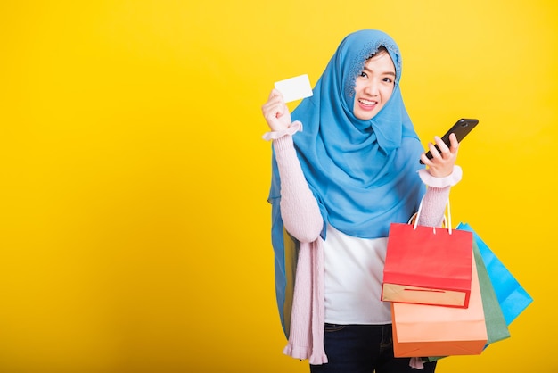 Árabe musulmán asiático, Retrato de una joven feliz y hermosa, religiosa islámica, con velo hijab, sonrisa graciosa, usando un teléfono inteligente en la mano y sosteniendo bolsas de compras, toma de estudio aislada en un fondo amarillo