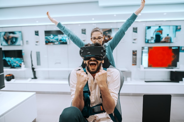 Raça mista atraente barbudo homem tentando a tecnologia de realidade virtual enquanto mulher assistindo o que ele está fazendo. Interior da loja de tecnologia.