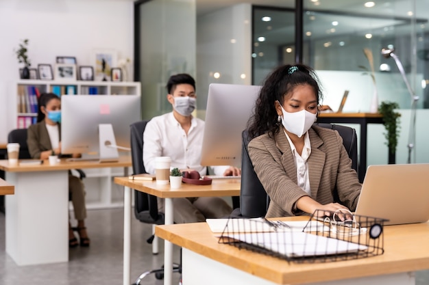 Raça mista de negra africana e mulher de negócios asiática usa máscara facial trabalhando em um novo escritório normal com distância social para Grupo de pessoas da equipe de negócios para prevenir a propagação do coronavírus COVID-19