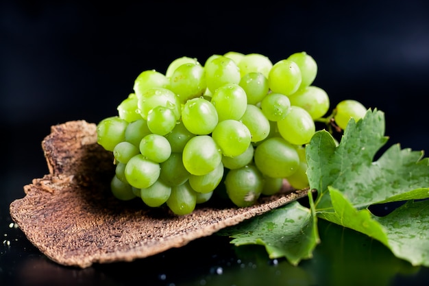 Racimo de uvas blancas jugosas con una hoja sobre un sustrato de madera y sobre un fondo negro