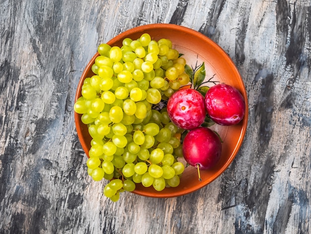 Racimo de uvas kishmouse verdes y ciruelas rojas en una placa de arcilla vista superior cerrar