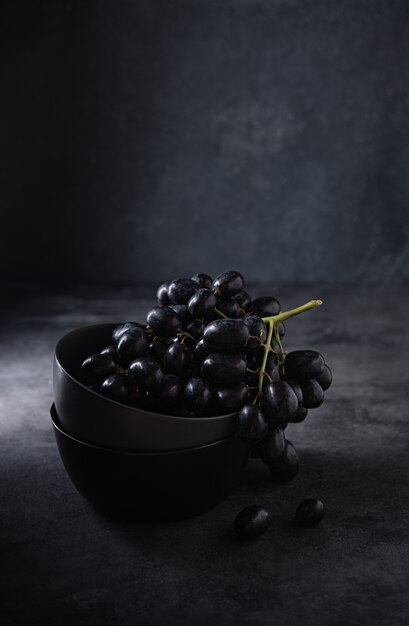 Un racimo de uvas negras jugosas yace en un recipiente gris oscuro sobre una mesa gris oscuro