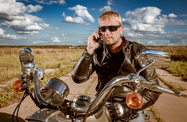 Radfahrer, der auf einem Smartphone spricht. Biker-Mann mit Lederjacke und Sonnenbrille sitzt auf seinem Motorrad.