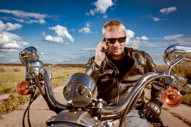 Radfahrer, der auf einem Smartphone spricht. Biker-Mann mit Lederjacke und Sonnenbrille sitzt auf seinem Motorrad.