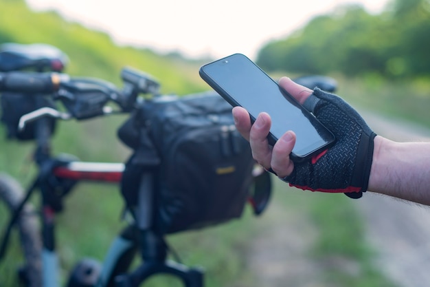 Radfahrer hält eine Smartphone-Nahaufnahme in den Händen vor dem Hintergrund eines Fahrrads in der Natur.