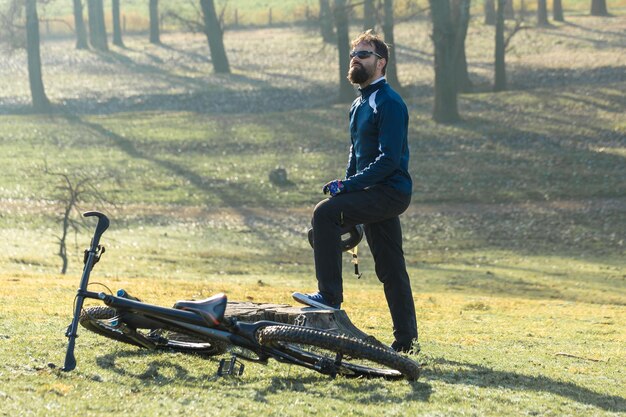 Radfahrer in Hose und Fleecejacke auf einem modernen Carbon-Hardtail-Bike mit Luftfedergabel fährt Offroad Der Typ ruht sich auf einem Baumstumpf im Park aus und blickt in die Ferne