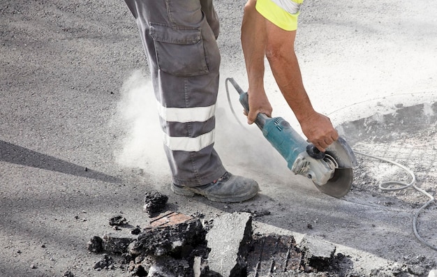 Foto radial com disco para pedra nas mãos de um operador de uniforme cortando o solo asfáltico, poeira i