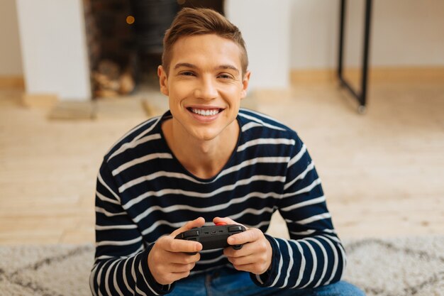 Radiante. Contenido atractivo joven rubio sonriendo y sosteniendo un control remoto para juegos mientras está sentado en el piso