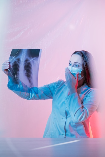 Radiologische Untersuchung Lungenpneumonie Coronavirus-Diagnose Infektion der Atemwege Verängstigte, schockierte Ärztin in Gesichtsmaske, die den Röntgenfilm der Brust in rosa Neonlicht untersucht
