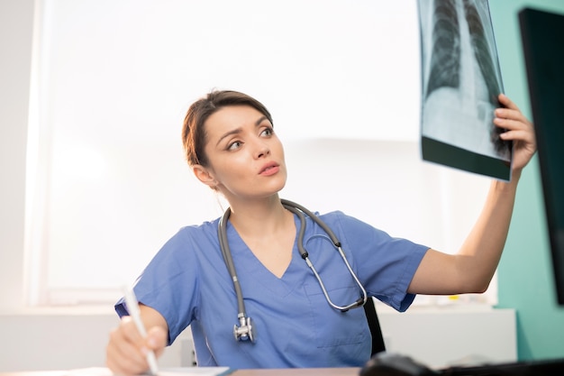 Radiólogo mujer morena joven mirando el resultado de rayos X de uno de los pacientes y haciendo notas de trabajo en clínicas