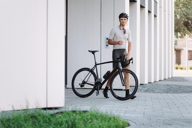 Radprofi in Sportoutfit und Brille posiert mit seinem schwarzen Fahrrad in der Nähe eines modernen Gebäudes Kaukasischer Mann verbringt Freizeit für aktives Training im Freien