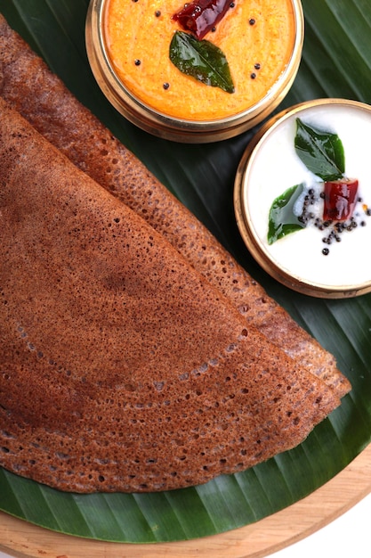 Foto ragi dosa, ein gesundes südindisches frühstücksprodukt, das auf einem runden holzsockel arrangiert ist, der mit bananenblättern und daneben platziertem kokosnuss-chutney ausgekleidet ist.