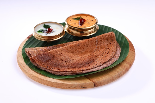 Foto ragi dosa, saludable desayuno del sur de la india dispuesto sobre una base de madera redonda forrada con hoja de plátano y chutney de coco colocada a su lado.