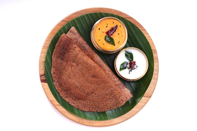 Ragi Dosa, saludable desayuno del sur de la India dispuesto sobre una base de madera redonda forrada con hoja de plátano y chutney de coco colocada a su lado.