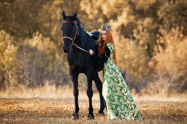 Rainha com cabelo ruivo em um vestido verde com uma coroa e perto de um cavalo