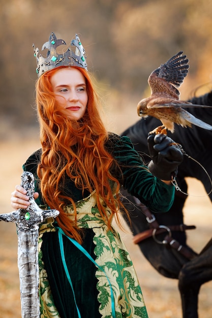 Foto rainha com cabelo ruivo em um vestido verde com uma coroa e uma espada perto de um cavalo com um pássaro