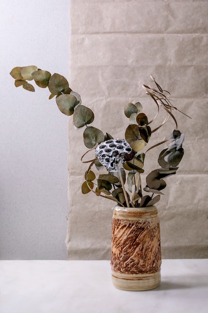 Rama de flores secas, eucalipto y ramitas en jarrón de cerámica marrón sobre mesa de mármol blanco con pared gris detrás.
