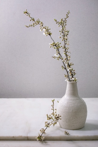 Ramas de cerezo florecientes en jarrón de porcelana blanca artesanal sobre mesa de mármol blanco.