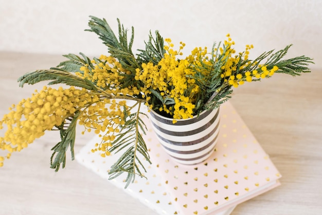 Ramas de flores de mimosa se encuentran en un jarrón de rayas blancas y negras en cuadernos sobre la mesa