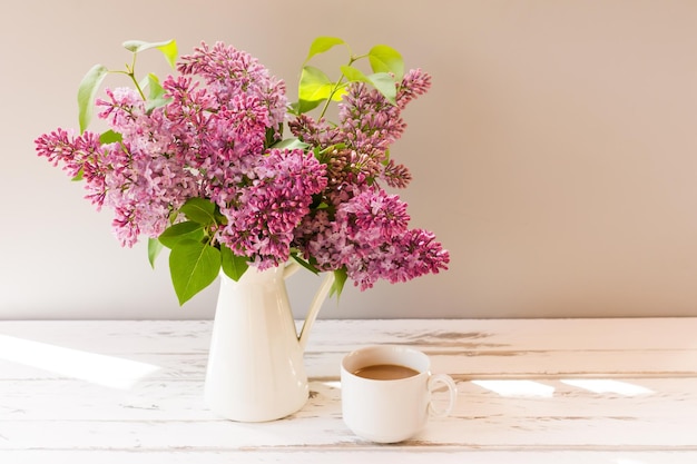 Ramas frescas de lilas en una jarra sobre una mesa de madera blanca con una taza de café a la sombra.