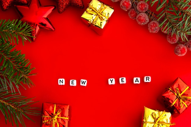 Ramas de pino verde con juguetes de Navidad rojos, cajas de regalo, bayas heladas rojas y la inscripción Año Nuevo en cubos sobre un fondo rojo.