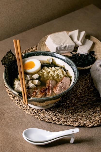 Ramen de sopa de estilo japonés casero con fideos tofu de pechuga de pato a la parrilla chips de nori de algas y huevo cocido en tazón de cerámica con palillos y cuchara en mesa marrón con servilleta de paja tejida