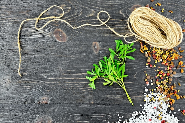 Raminho de arruda verde fresca com sal, pimenta, sementes de feno-grego e um rolo de barbante contra uma placa de madeira preta