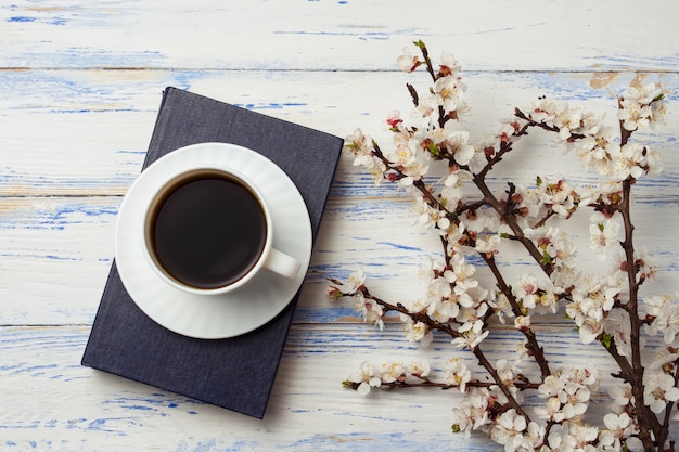 Raminho de cerejas com flores e copo branco com café preto e um livro sobre um fundo branco de madeira.