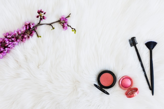 Ramita de flor morada con polvos faciales compactos y pinceles de maquillaje sobre fondo de piel