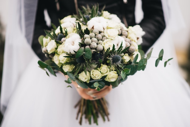 Ramo de la boda en manos de la novia. Ramo Khimki con rosas blancas, algodón, espinas y vegetación.