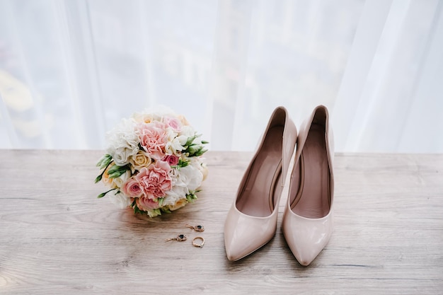 Ramo de bodas de la novia de flores rosadas rosas y vegetación elegante clásico lacado beige zapatos arete y anillo sobre fondo pastel Accesorios de novia Primer plano vista superior