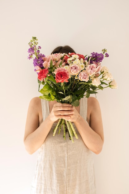 Ramo de flores de colores en manos femeninas sobre fondo blanco Mujer joven en vestido de lino lavado beige neutro ocultar cara Belleza estética y concepto floral de moda Rostro oculto por ramo de flores