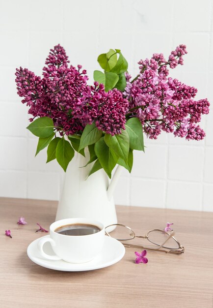 Ramo de flores lilas en un jarrón y una taza de café sobre una mesa de madera.