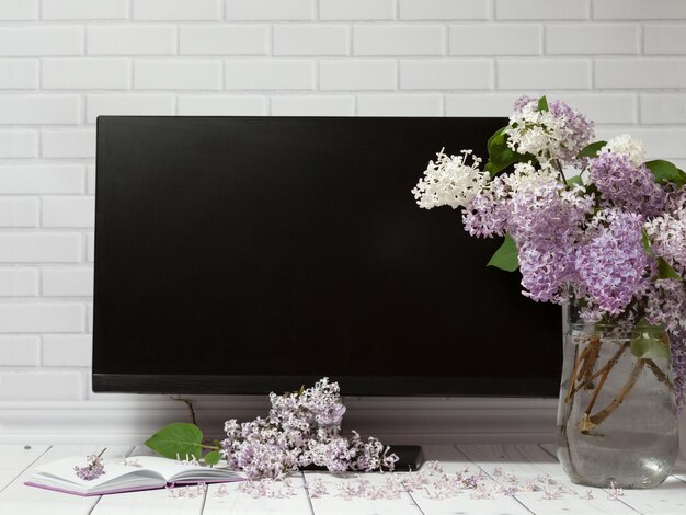 Ramo de flores lilas en jarrón de vidrio, cuaderno abierto, pantalla de monitor negra, espacio de trabajo de primavera