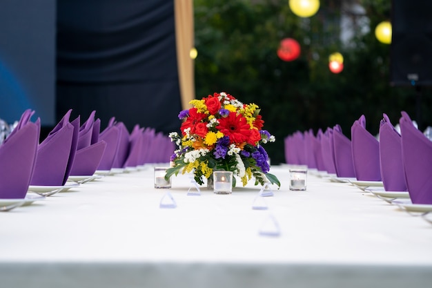 El ramo de flores rojo, amarillo, púrpura y blanco está colocado en la mesa de cubierta blanca larga y listo para una cena de lujo en el campo del jardín.