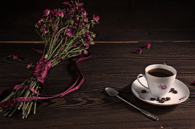 Ramo de flores secas con una cinta sobre la superficie de madera oscura. Una taza de café de porcelana, una cuchara de plata y un platillo.